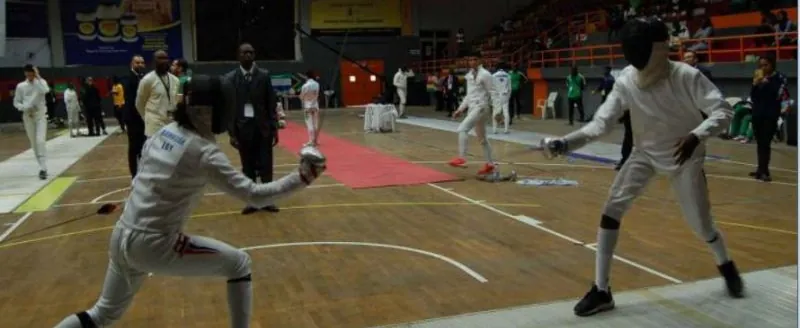 Two fencers in white kit competing on the piste at a championship in Nigeria, with officials watching