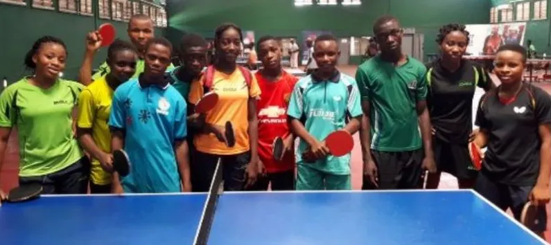 Young Nigerian table tennis players in club shirts hold their bats beside a blue table at a training camp